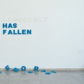 Wall with hung letters forming the words "Has Fallen". On the floor are the letters for the word "Democracy"