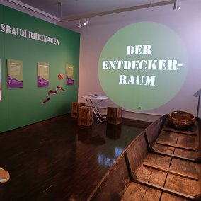 Blick in die Ausstellung, © Maria Karipidou/Stadtmuseum zu sehen sind Infotafeln im Museumsraum mit Sitzgelegenheit, dem Nachen und im Vordergrund die Maus einkopiert